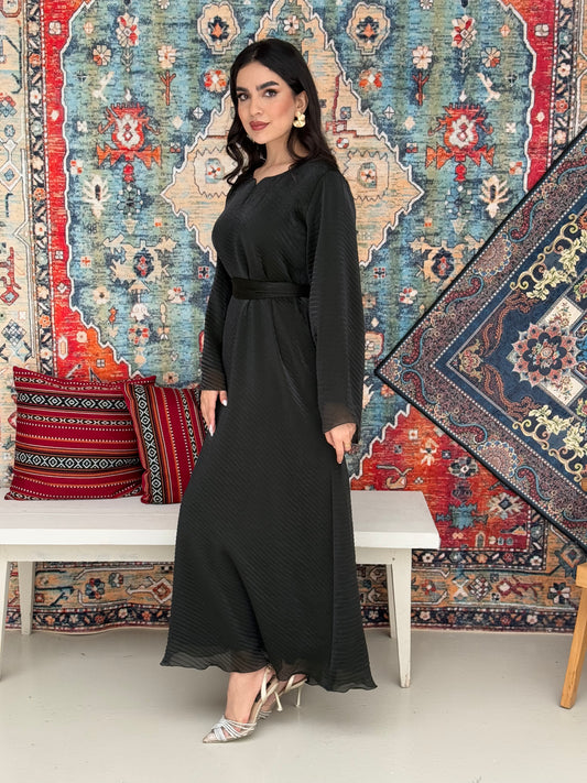 A woman standing in front of a decorative rug, wearing a Dark Green Shimmer Organza dress with long sleeves and a belt, with her hair styled in an updo.