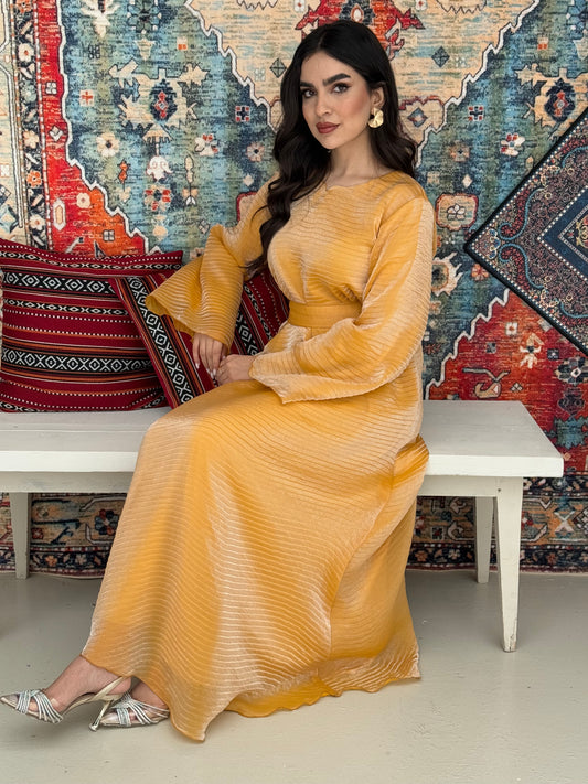 A woman standing in front of a decorative rug, wearing a golden yellow Shimmer Organza dress with long sleeves and a belt, with her hair styled in an updo.