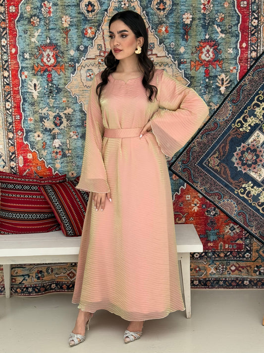 A woman standing in front of a decorative rug, wearing a peach gold Shimmer Organza dress with long sleeves and a belt, with her hair styled in an updo.