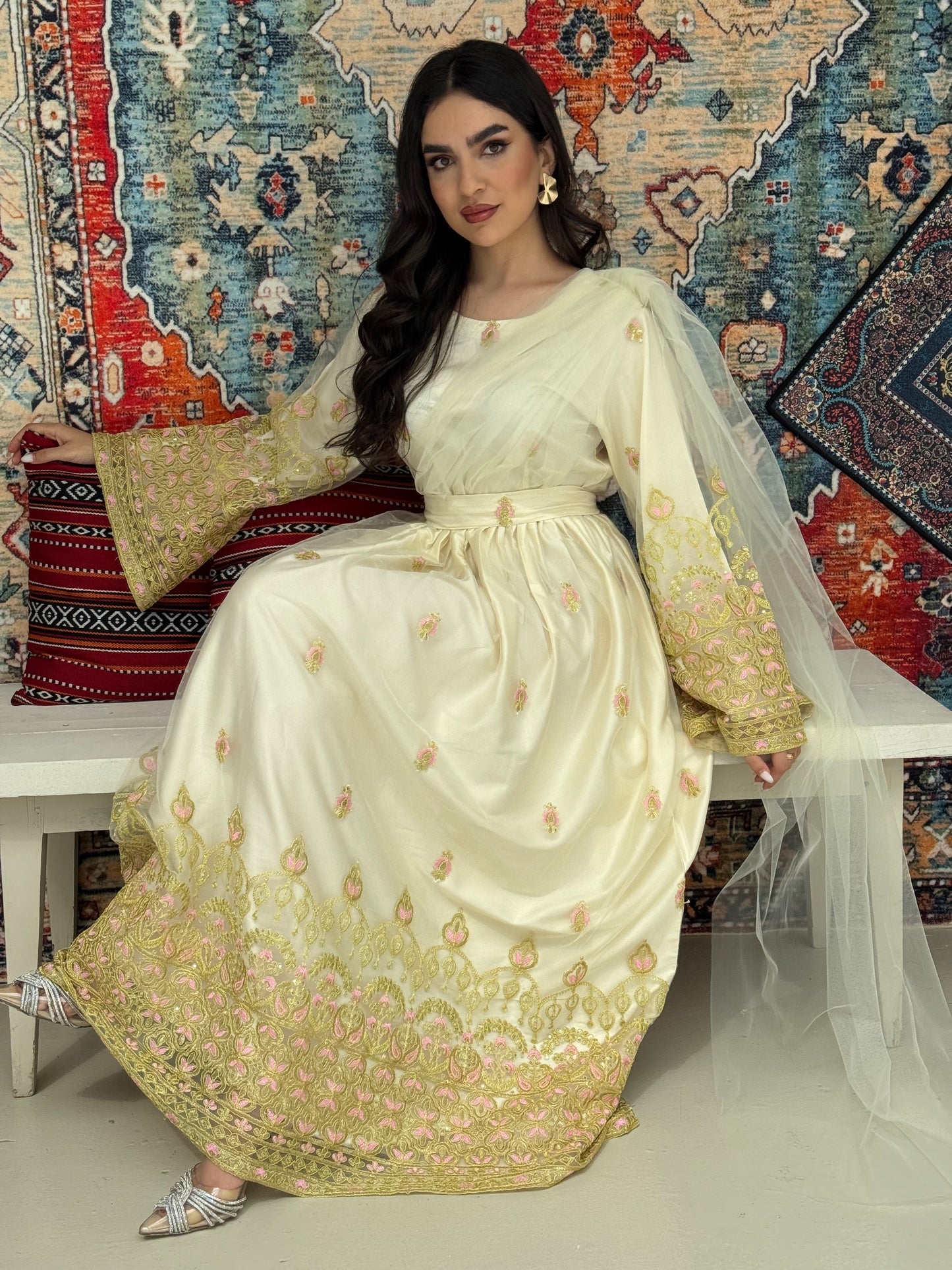 A woman sitting in front of a colorful backdrop wearing a cream and gold-colored Saree-Style Draped Jalabiya with net embroidery, featuring a belt.