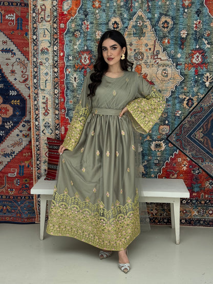 A woman standing in front of a colorful backdrop wearing a sage green and gold-colored Saree-Style Draped Jalabiya with net embroidery, featuring a belt.