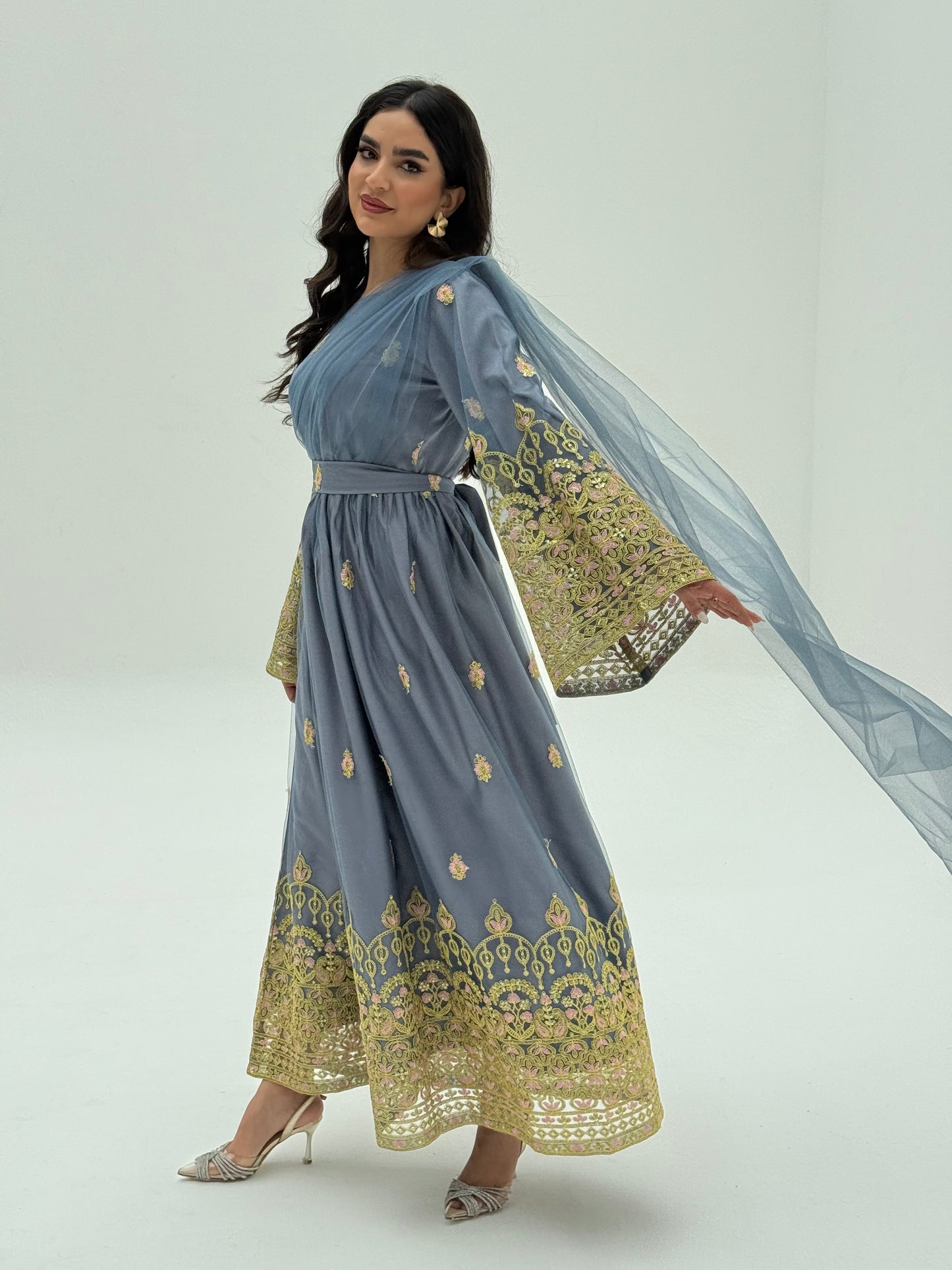 A woman standing in front of a colorful backdrop wearing a Steel Blue and gold-colored Saree-Style Draped Jalabiya with net embroidery, featuring a belt.