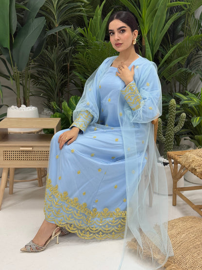 A woman seated in a tropical setting, wearing a baby blue jalabiya dress with gold embroidery and a belt, posing with a smile.