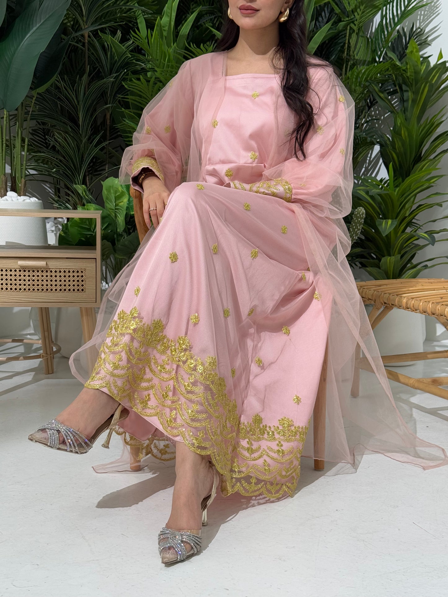 A woman seated in a tropical setting, wearing a baby pink jalabiya dress with gold embroidery and a belt, posing with a smile.