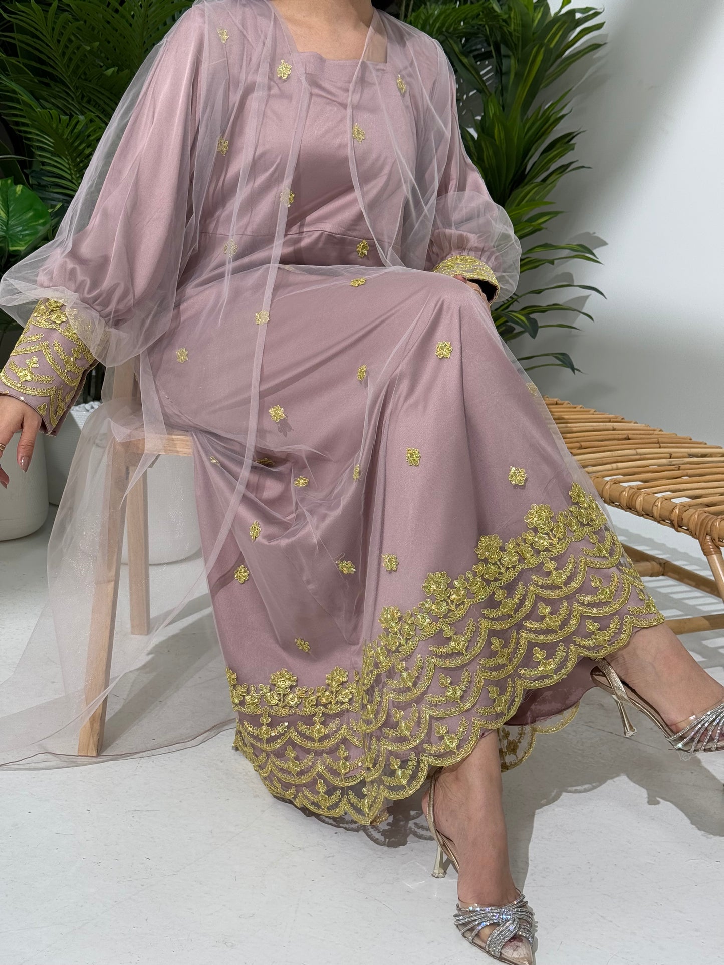 A woman seated in a tropical setting, wearing a light purple jalabiya dress with gold embroidery and a belt, posing with a smile.