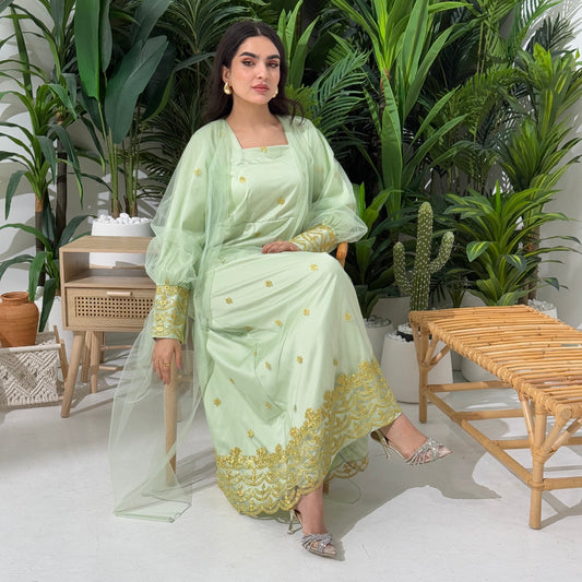 A woman seated in a tropical setting, wearing a green dress jalabiya with gold embroidery and a belt, posing with a smile.