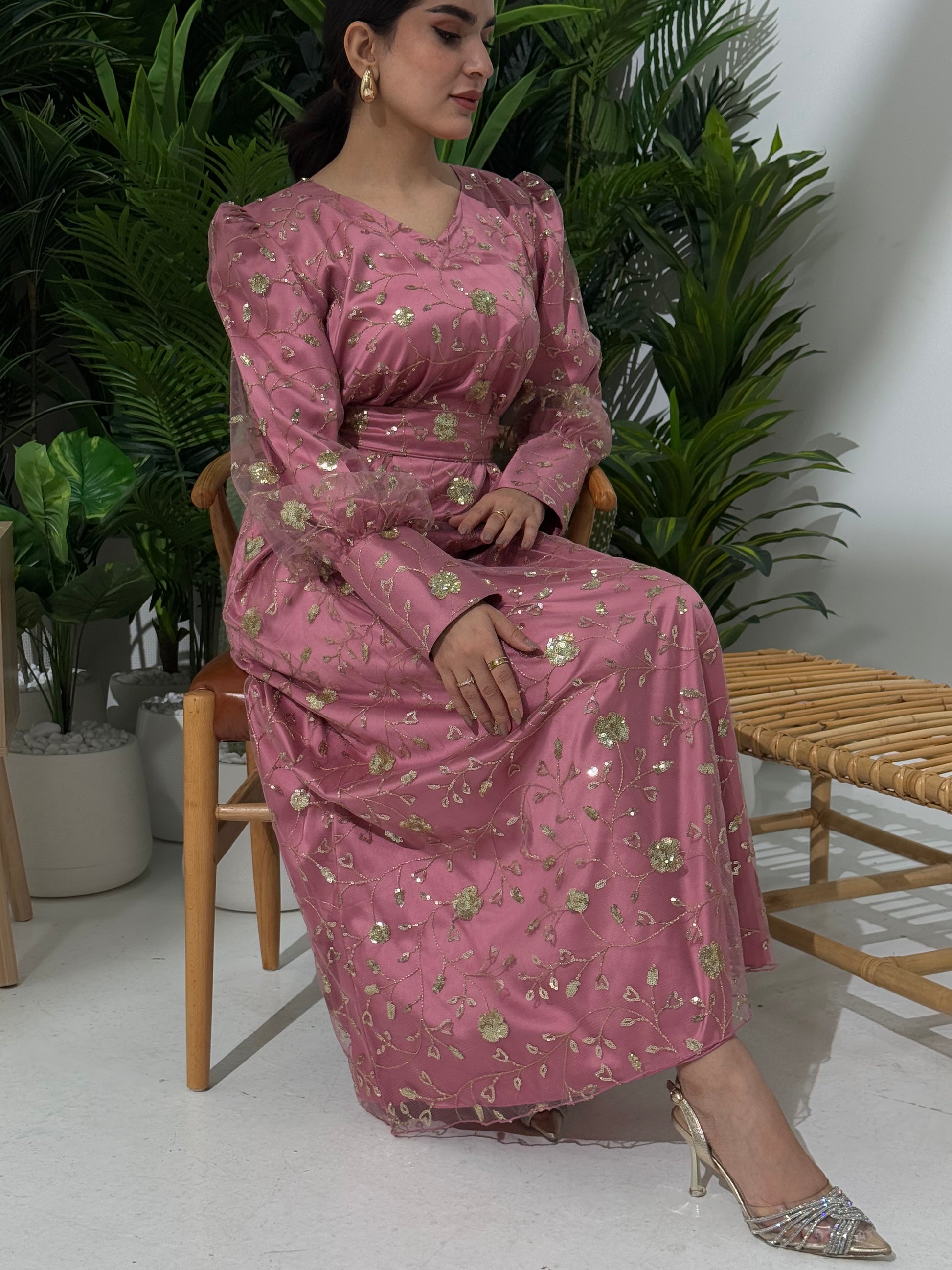 A woman sitting in a tropical setting wearing a dusty rose  Sheer Net Overlay with gold Floral Embroidery dress and a belt.