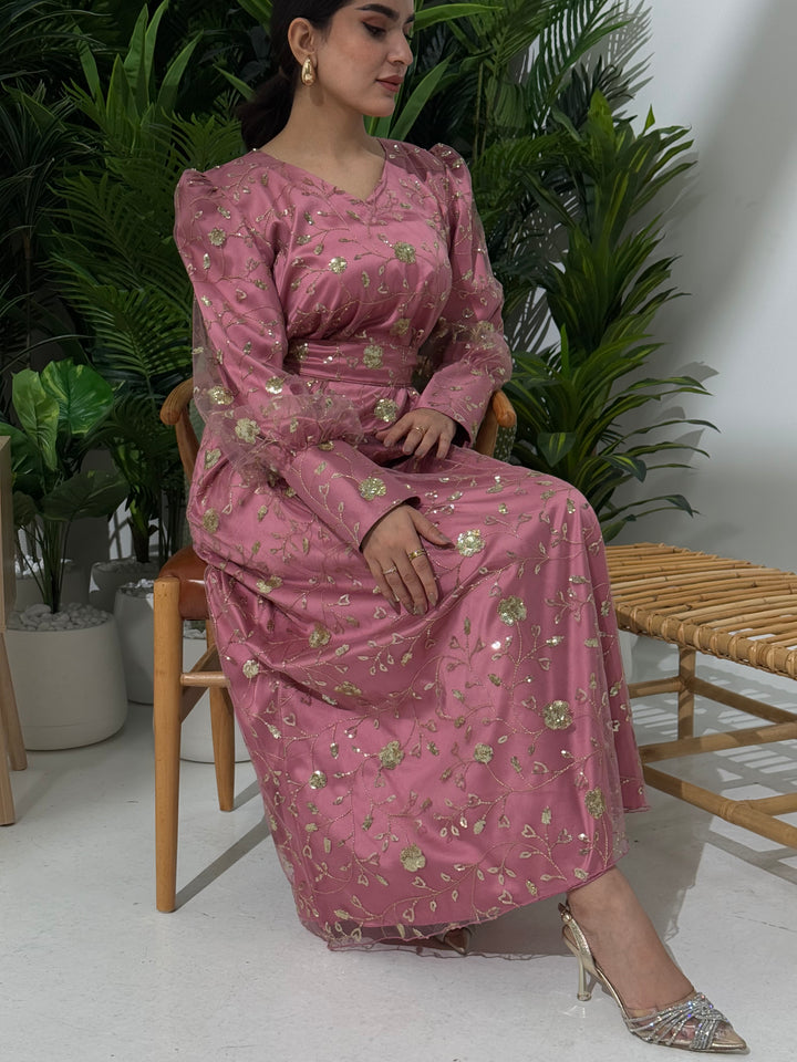 A woman sitting in a tropical setting wearing a dusty rose  Sheer Net Overlay with gold Floral Embroidery dress and a belt.