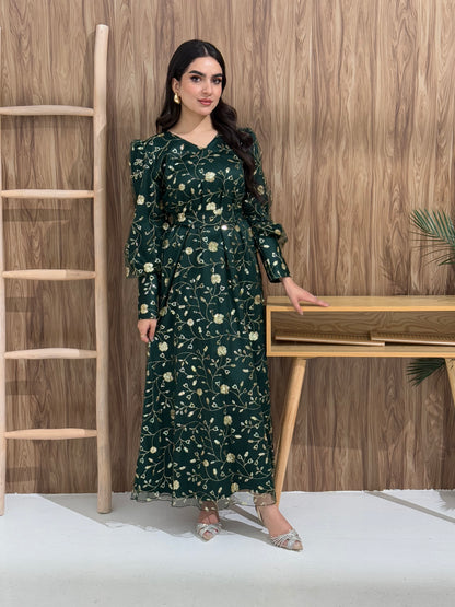 A woman sitting in a tropical setting wearing an emerald green Sheer Net Overlay with gold Floral Embroidery dress and a belt.
