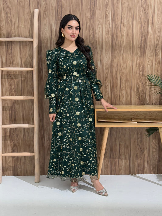 A woman sitting in a tropical setting wearing an emerald green Sheer Net Overlay with gold Floral Embroidery dress and a belt.