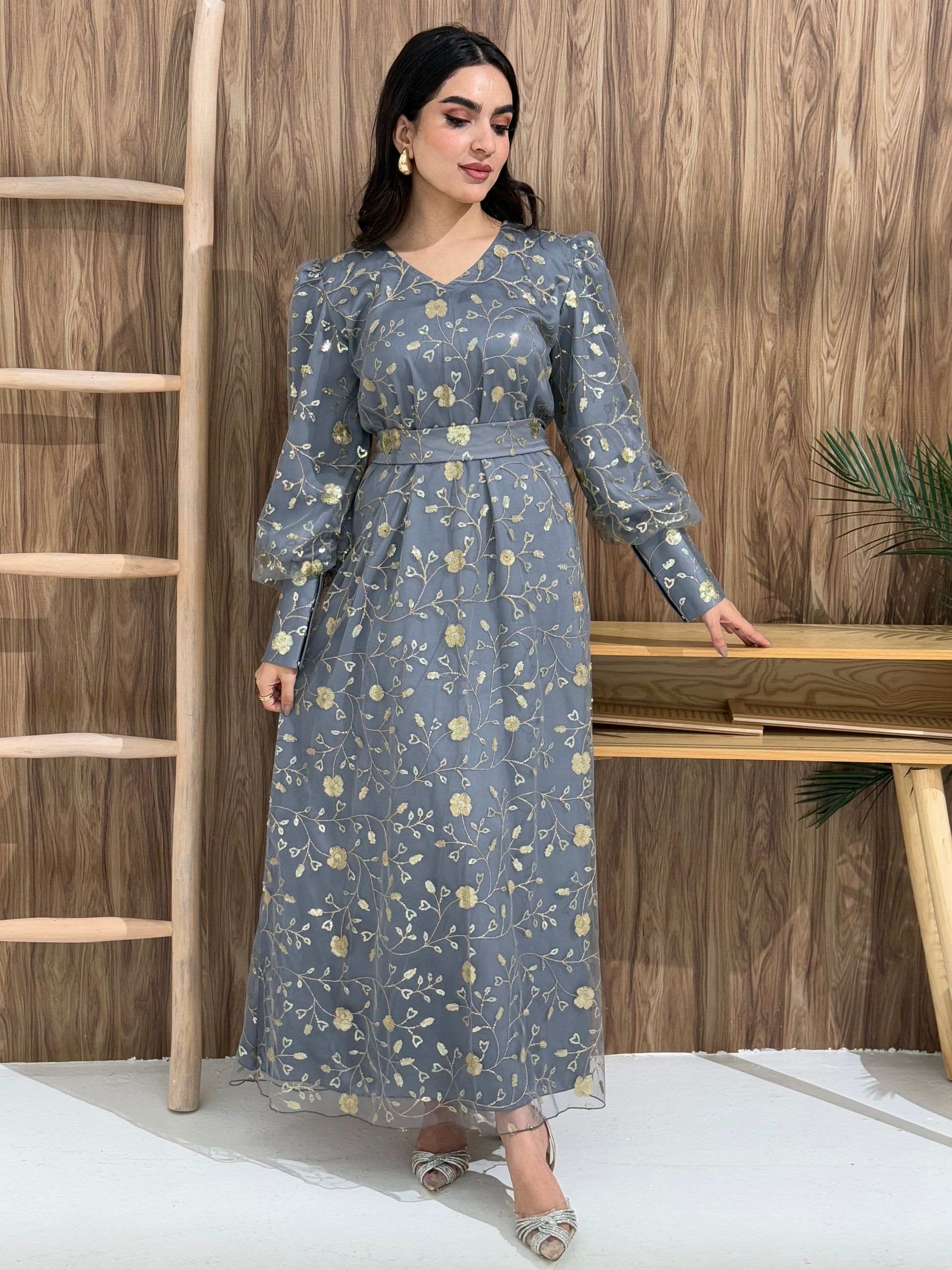 A woman sitting in a tropical setting wearing a grey Sheer Net Overlay with gold Floral Embroidery dress and a belt.