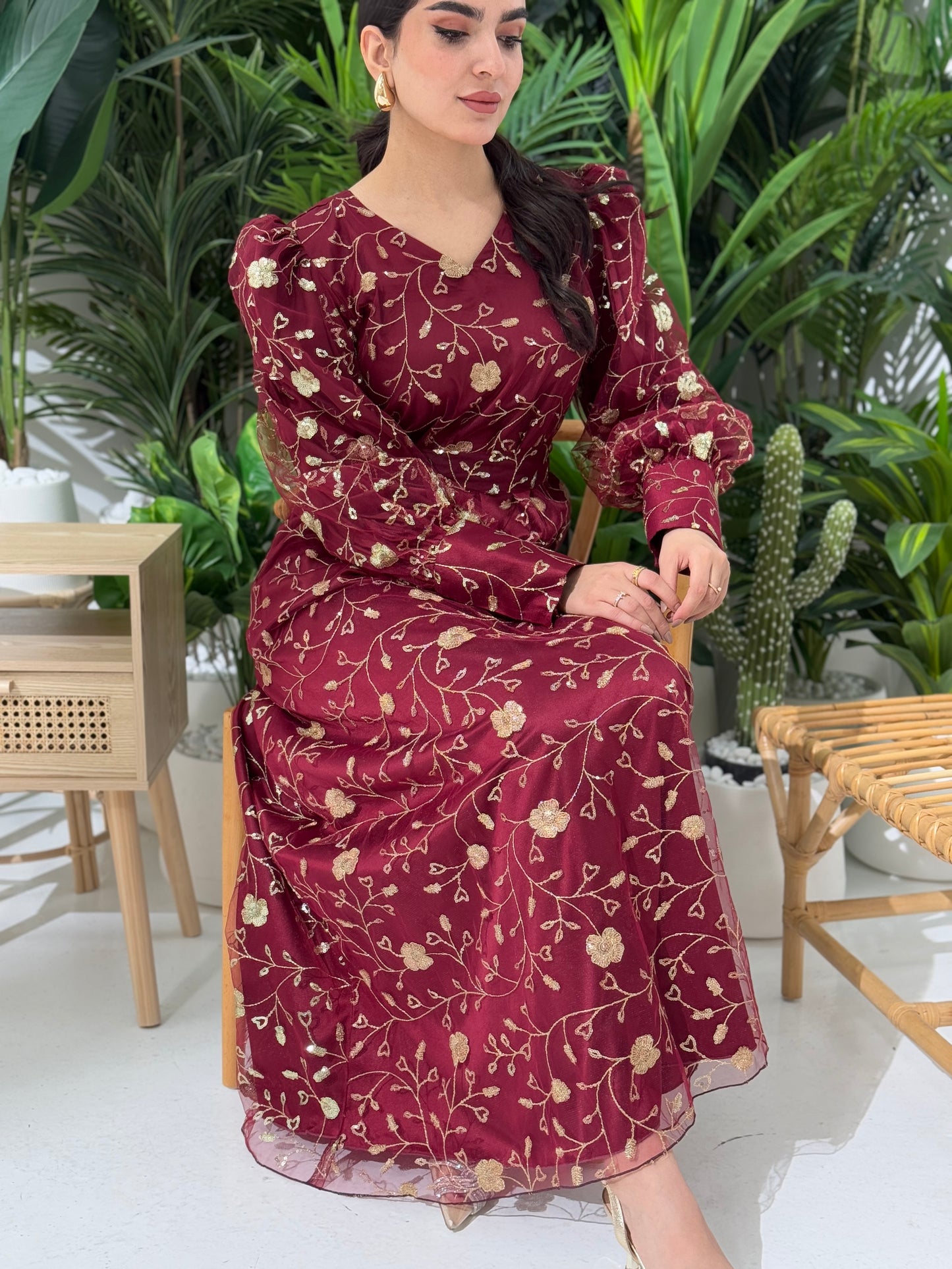 A woman sitting in a tropical setting wearing a maroon Sheer Net Overlay with gold Floral Embroidery dress and a belt.