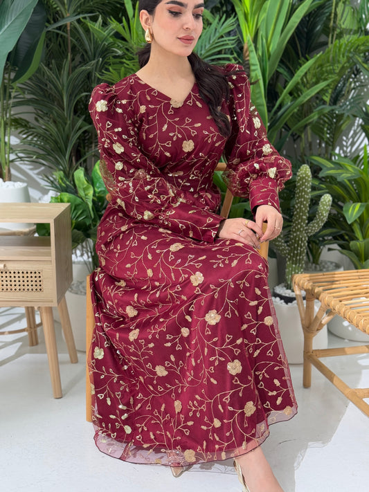 A woman sitting in a tropical setting wearing a maroon Sheer Net Overlay with gold Floral Embroidery dress and a belt.