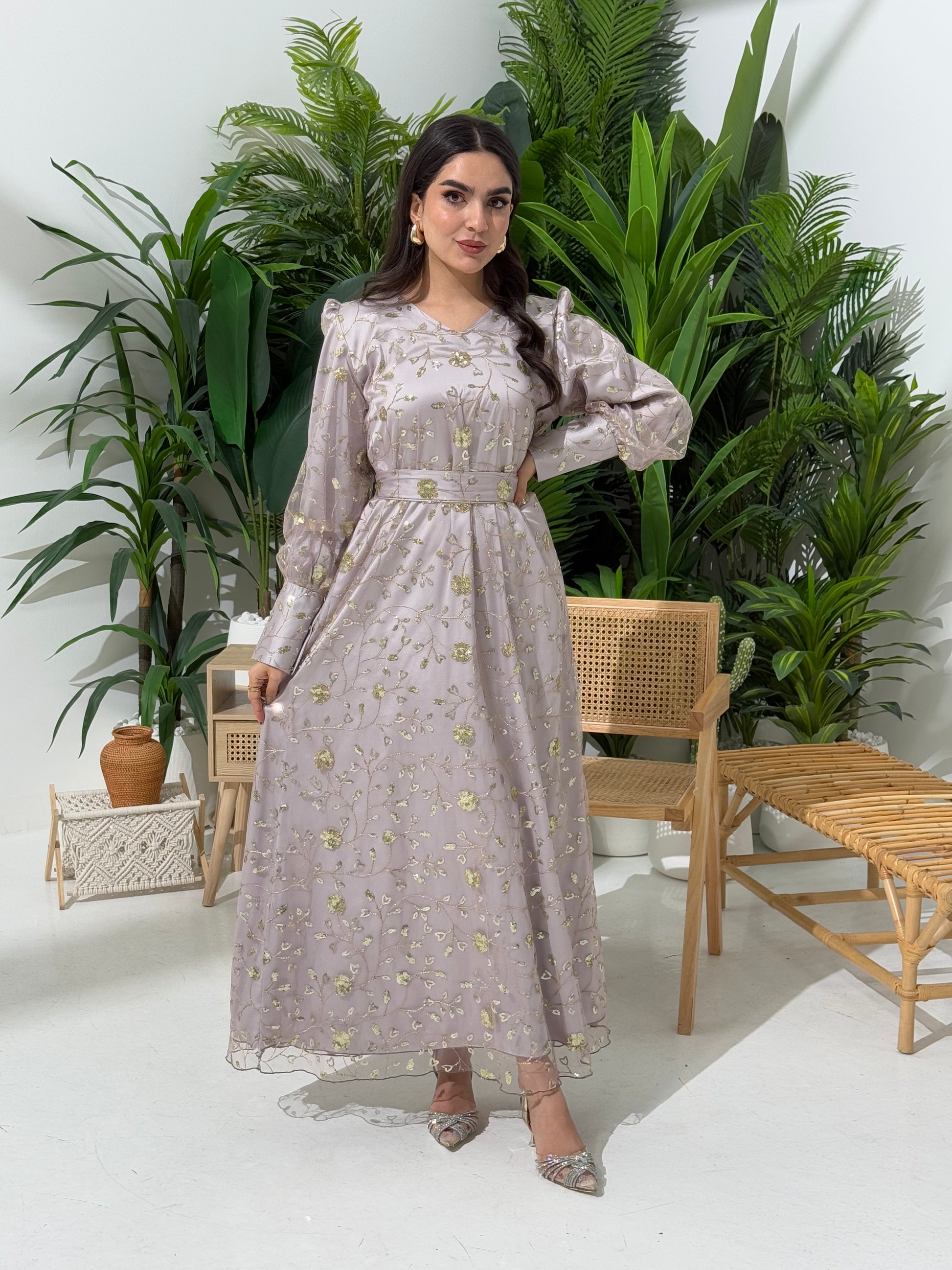 A woman standing in a tropical setting wearing a silver grey Sheer Net Overlay with gold Floral Embroidery dress and a belt.