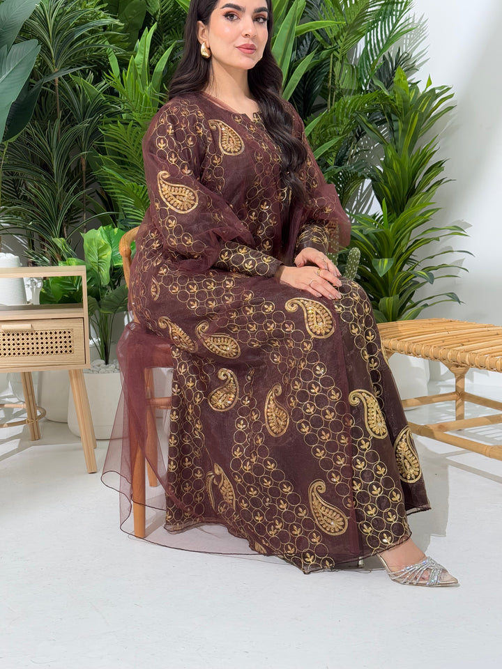 A woman seated in a garden setting, wearing an A-line brown Paisley Dress with Bishop Sleeves with gold embroidery and a netted veil.