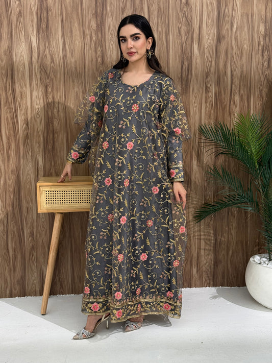 A woman standing in an indoor setting, wearing a dark grey dress with Pink floral embroidery, gold vine patterns, wide, draped sleeves, and a satin base with a Sheer Net Overlay.