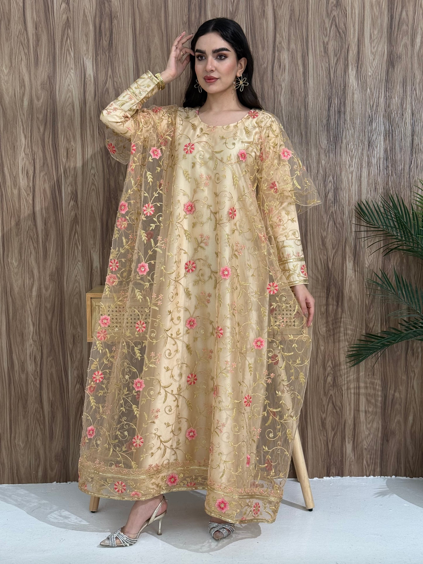 A woman standing in an indoor setting, wearing a gold beige dress with Pink floral embroidery, gold vine patterns, wide, draped sleeves, and a satin base with a Sheer Net Overlay.