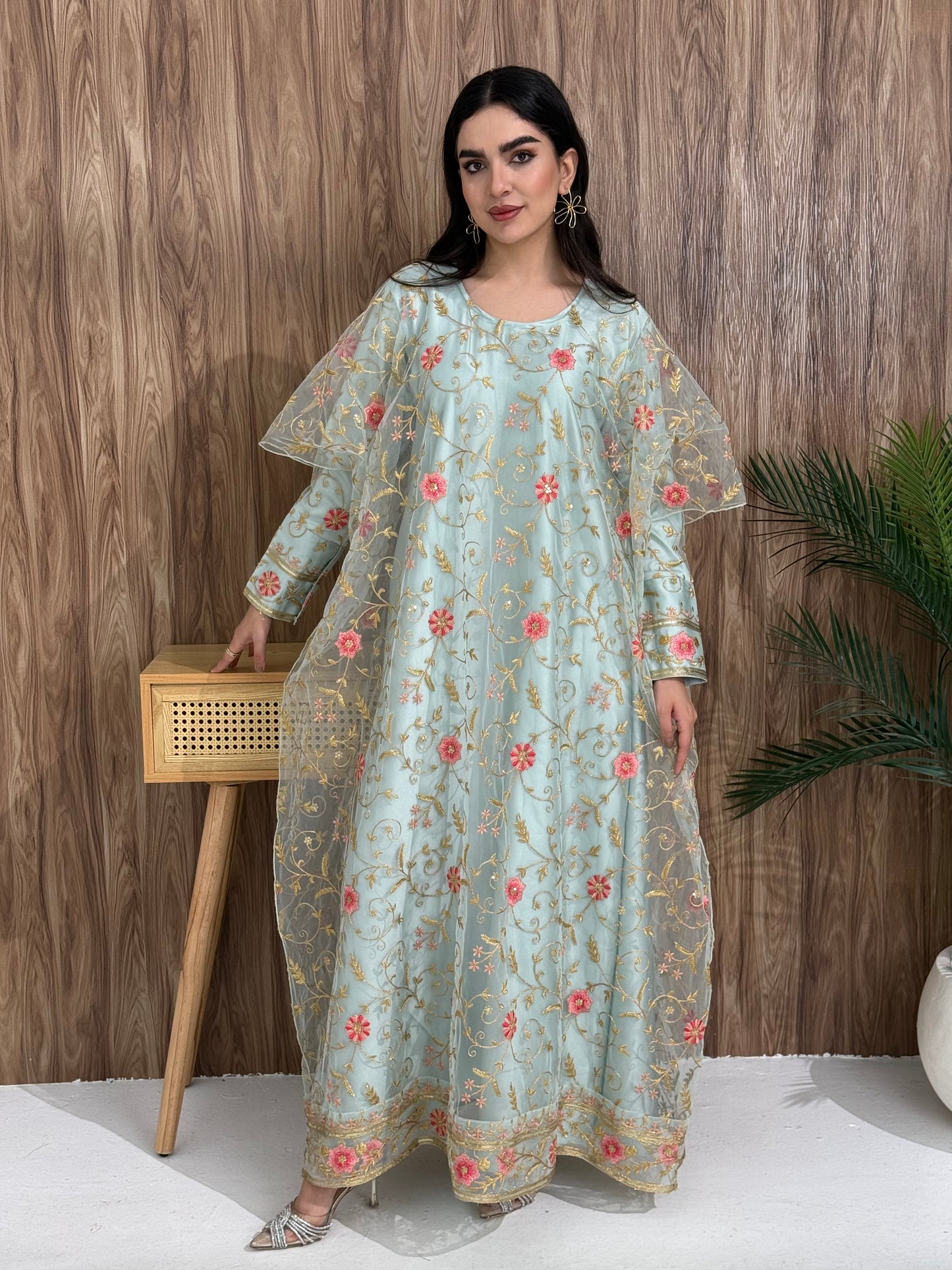 A woman standing in an indoor setting, wearing a light blue dress with Pink floral embroidery, gold vine patterns, wide, draped sleeves, and a satin base with a Sheer Net Overlay.