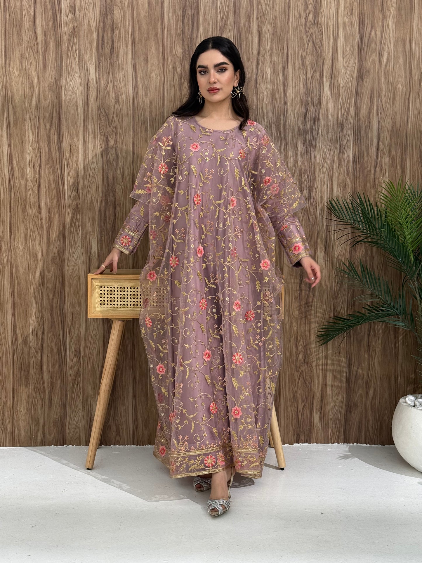 A woman standing in an indoor setting, wearing a light purple dress with Pink floral embroidery, gold vine patterns, wide, draped sleeves, and a satin base with a Sheer Net Overlay.