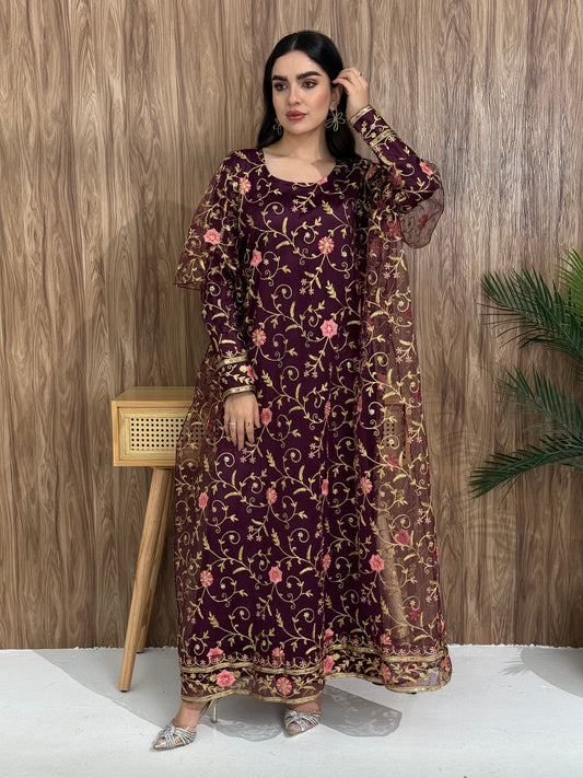 A woman standing in an indoor setting, wearing a maroon dress with Pink floral embroidery, gold vine patterns, wide, draped sleeves, and a satin base with a Sheer Net Overlay.
