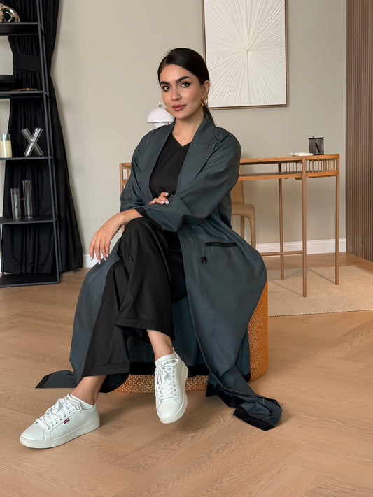 Woman sitting on a chair wearing a long dark teal Linen Abaya Set, white sneakers, and beige pants.