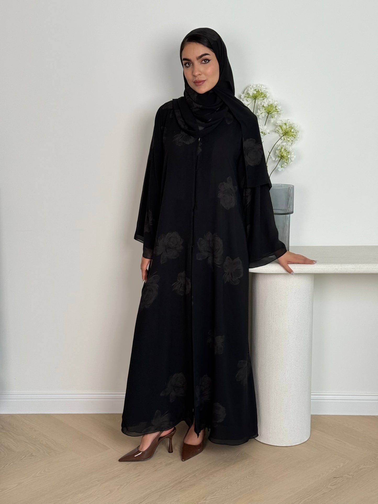 Woman wearing a Black & Beige abaya with floral patterns and a matching sheila in a minimalistic setting.