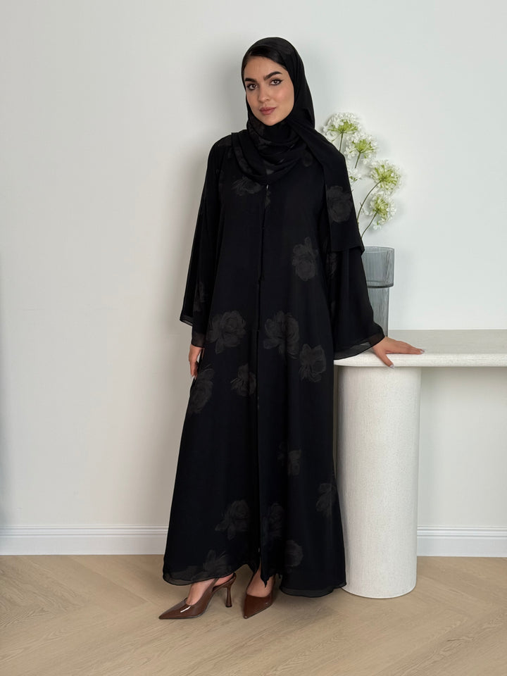 Woman wearing a Black & Beige abaya with floral patterns and a matching sheila in a minimalistic setting.