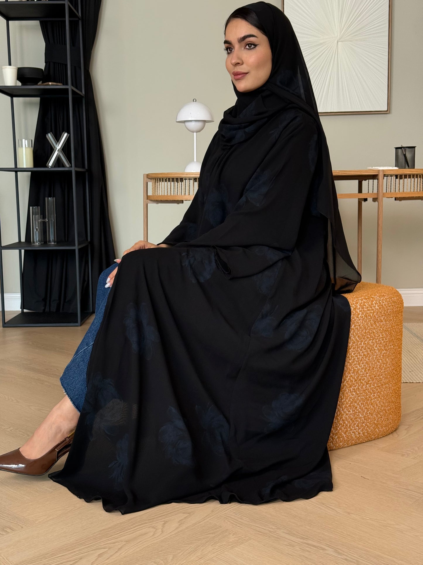 Woman wearing a Black & Midnight Blue Floral Satin Abaya and a matching sheila in a room with furniture and decor.