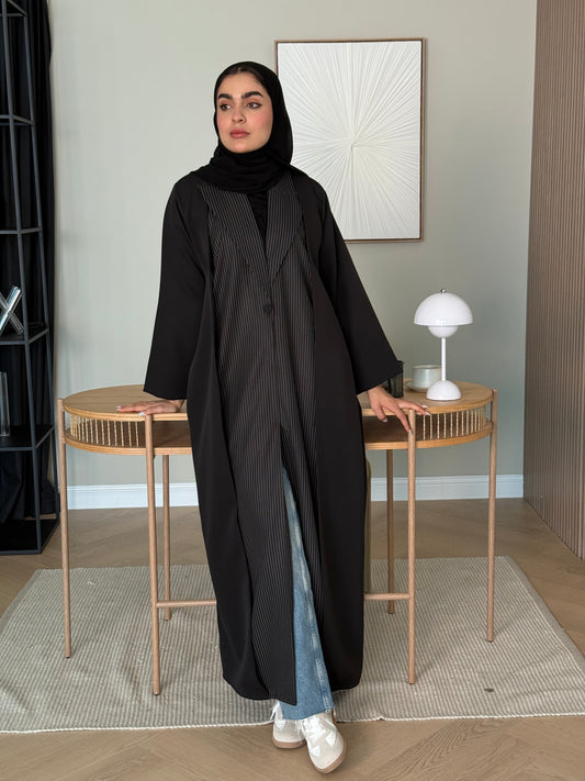 Woman in a black pinstripe linen abaya and matching sheila standing in a room with decor elements.
