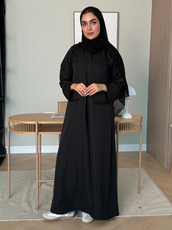 Person wearing a black linen pocket abaya  with matching sheila standing indoors with a neutral background
