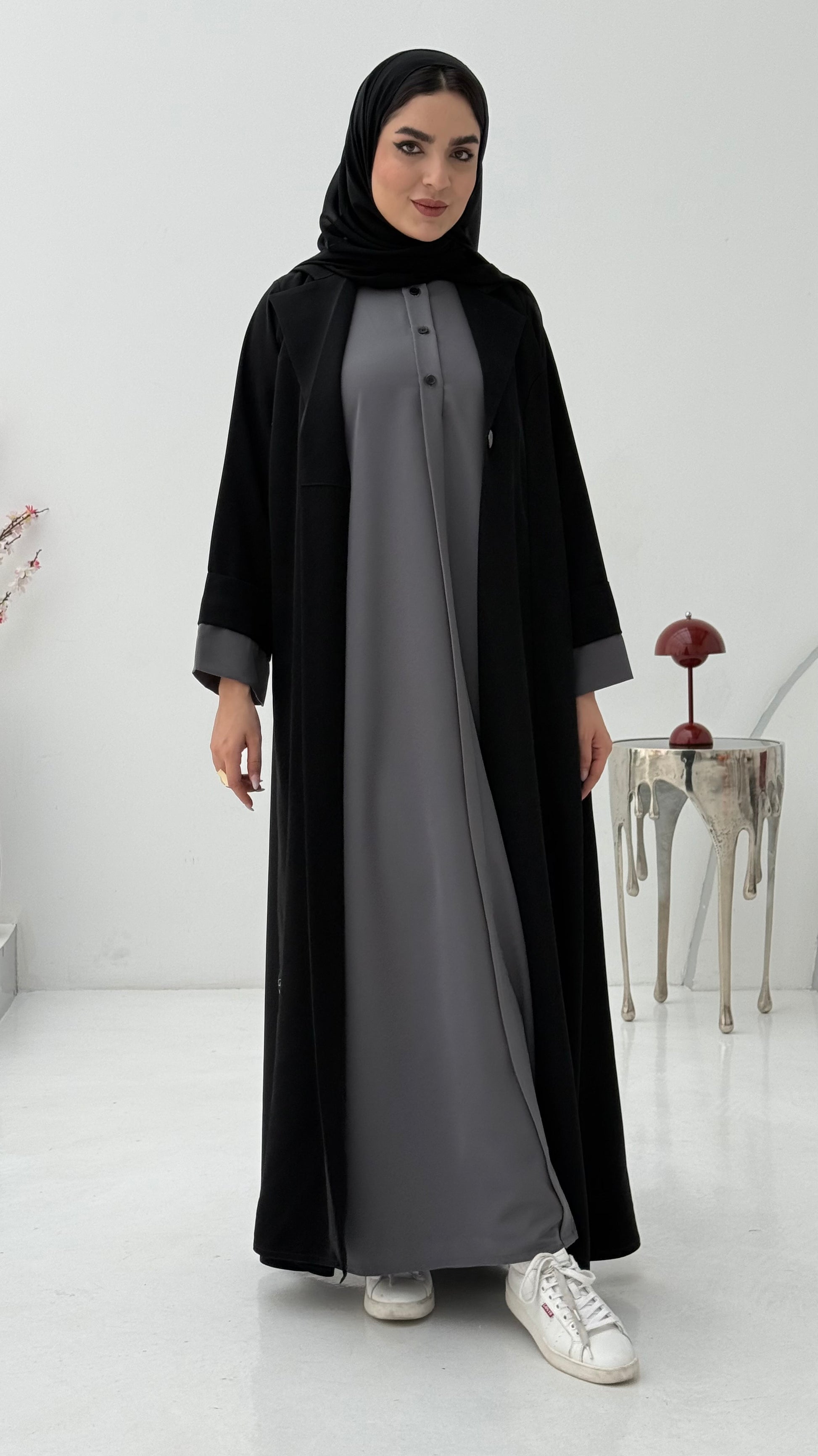 Woman wearing a black abaya over a gray dress and a matching sheila on a white background