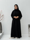 Woman wearing a black abaya and matching sheila in a minimalistic setting with a vase and decorative items.