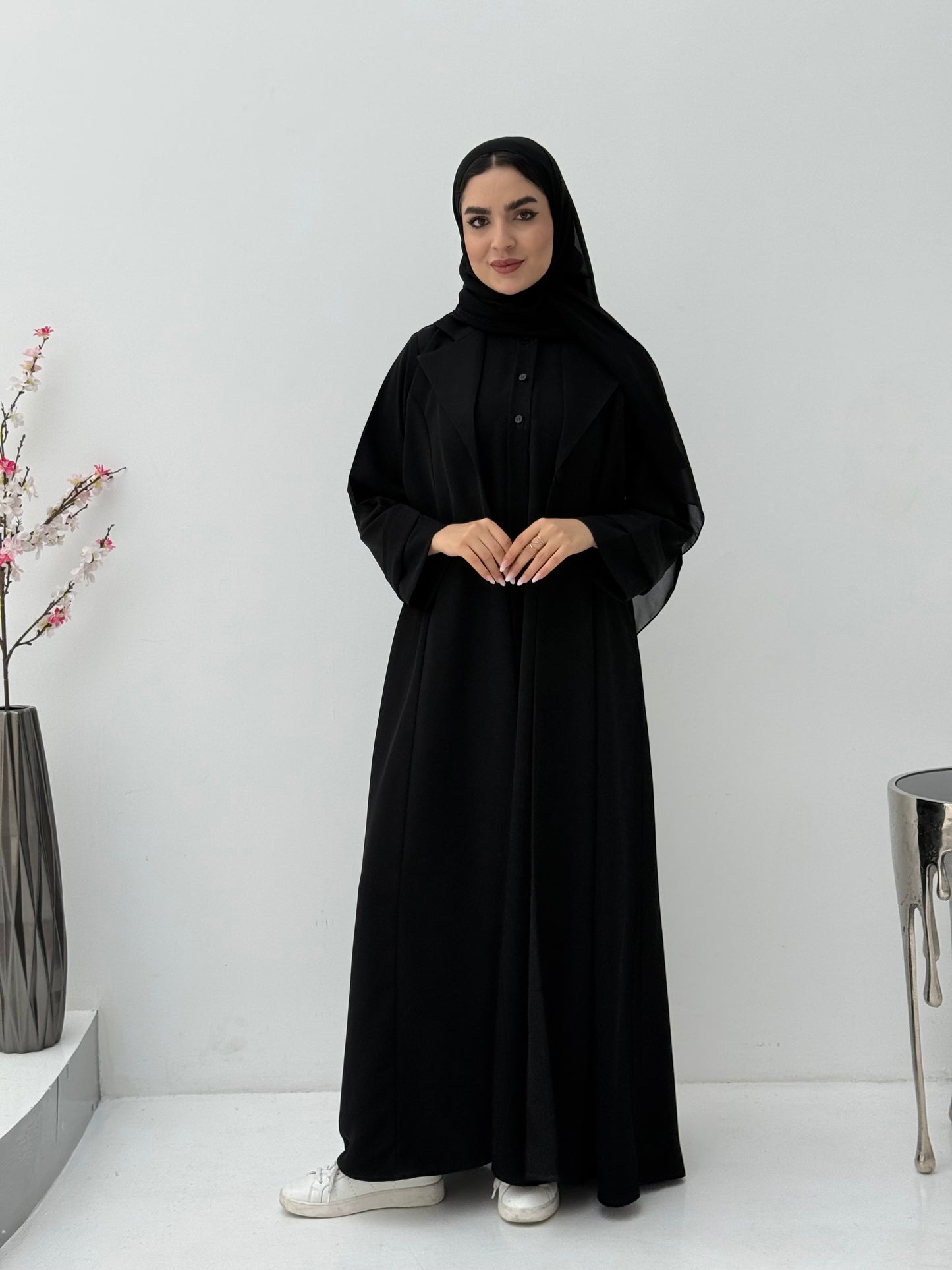 Woman wearing a black abaya and matching sheila in a minimalistic setting with a vase and decorative items.