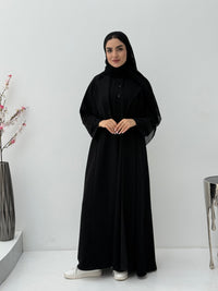 Woman wearing a black abaya and matching sheila in a minimalistic setting with a vase and decorative items.
