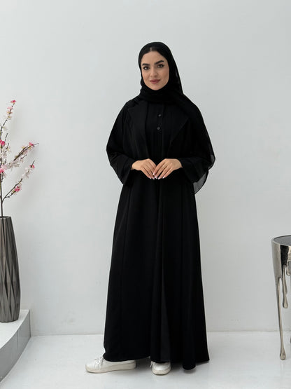 Woman wearing a black abaya and matching sheila in a minimalistic setting with a vase and decorative items.