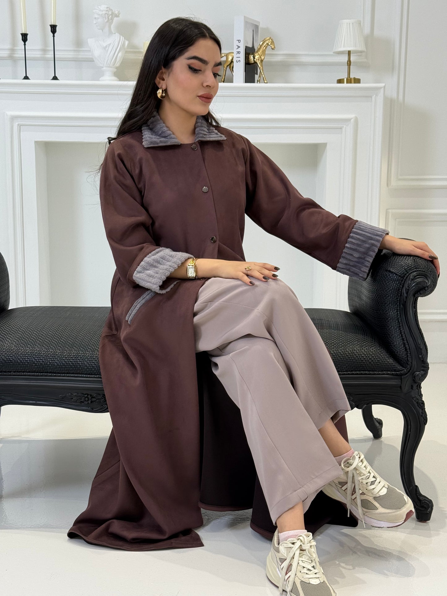 Woman in a Burgundy Shamwa Abaya and gray pants sitting on a chair with a white background
