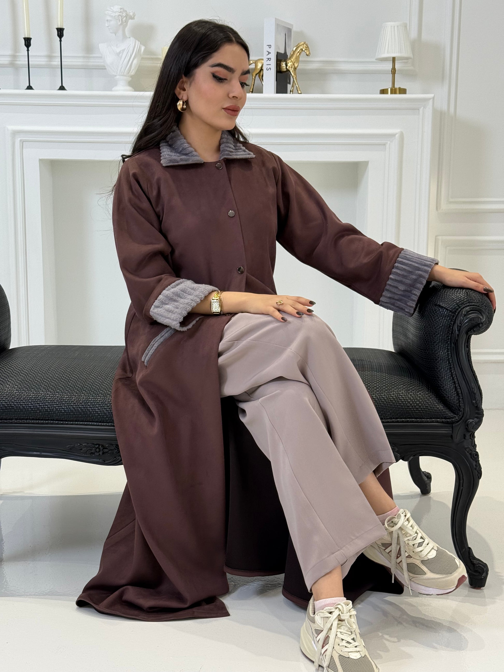 Woman in a Burgundy Shamwa Abaya and gray pants sitting on a chair with a white background