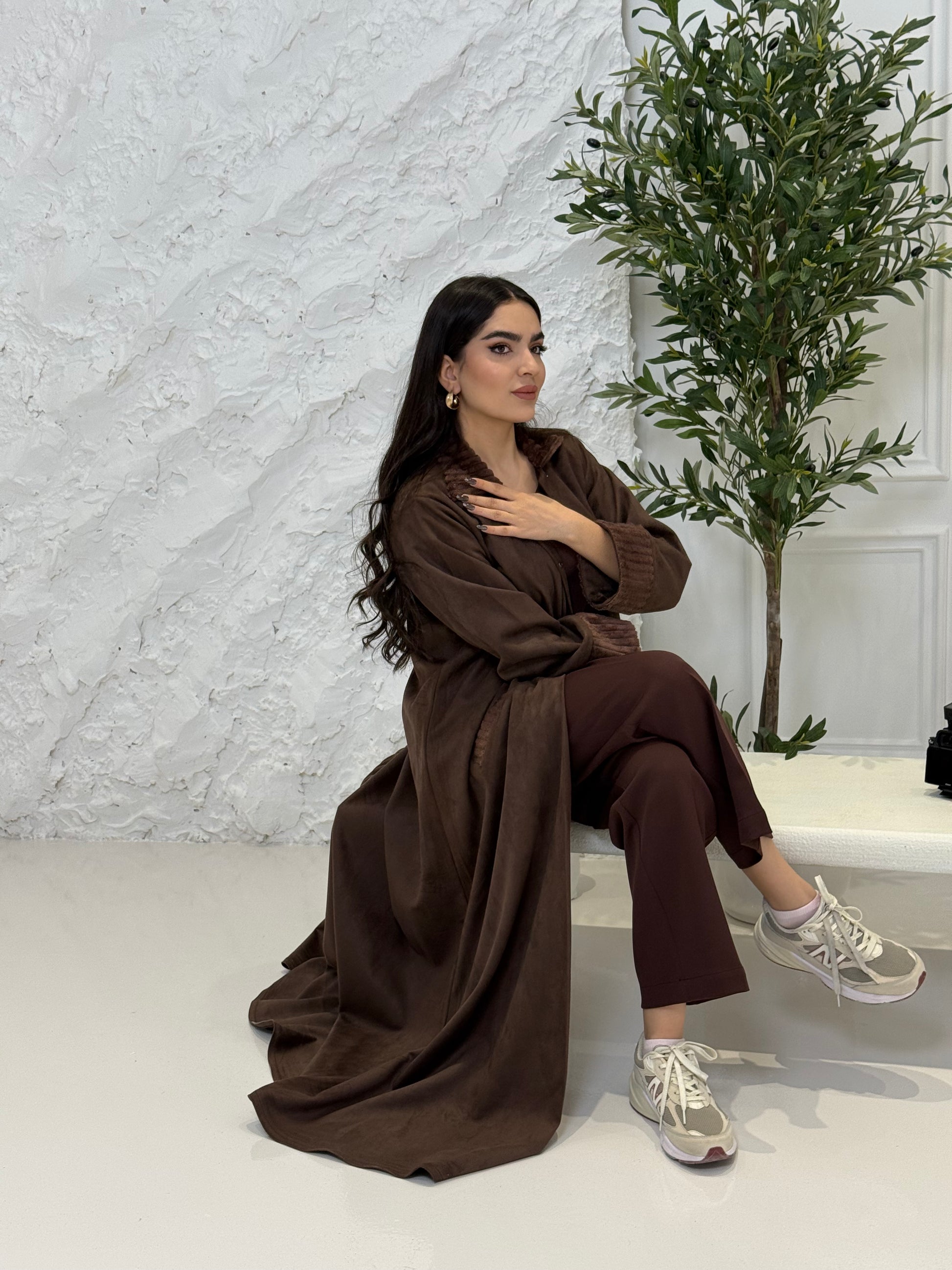 Woman in a Dark Brown Shamwa Abaya  sitting on a white carpet with a plant in the background