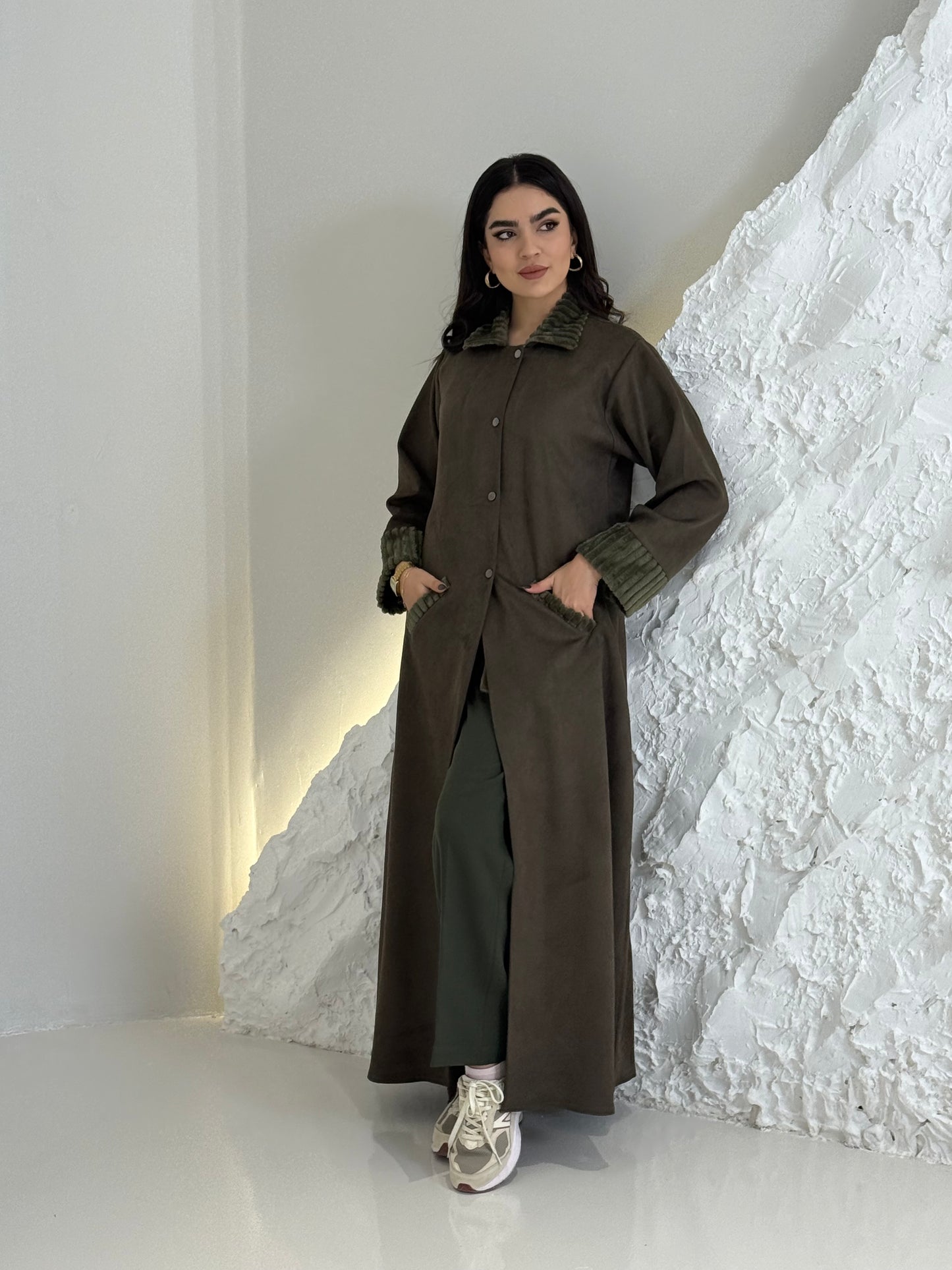 Woman wearing a long Dark Olive Shamwa Abaya Set standing against a textured white wall.