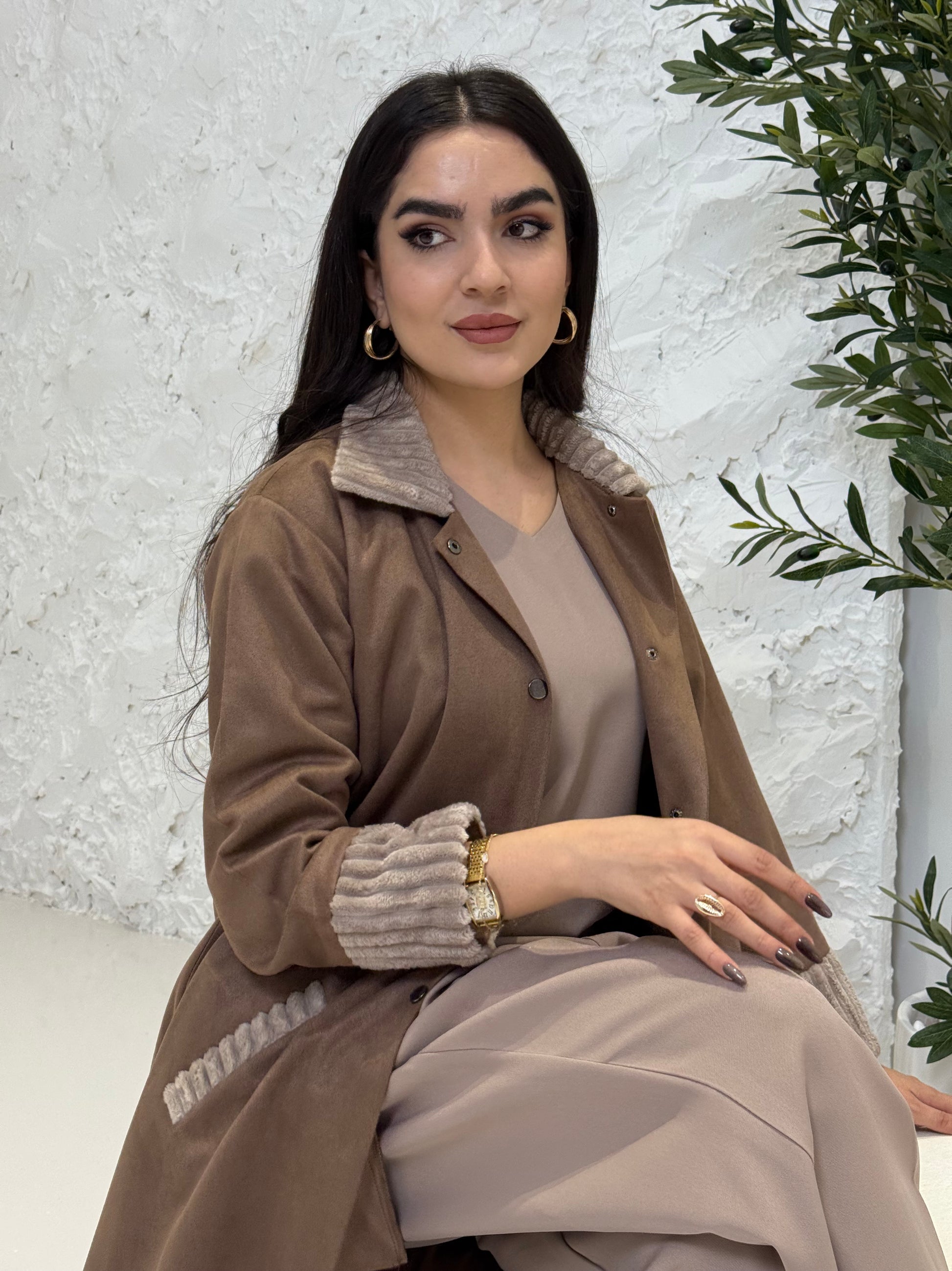 Woman in a Light Brown Shamwa Abaya brown coat standing on a textured white surface with greenery.