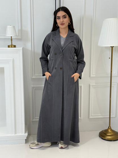 Woman wearing a  Dark Grey Shamwa Trench Abaya Set standing in a room with white walls and a lamp.