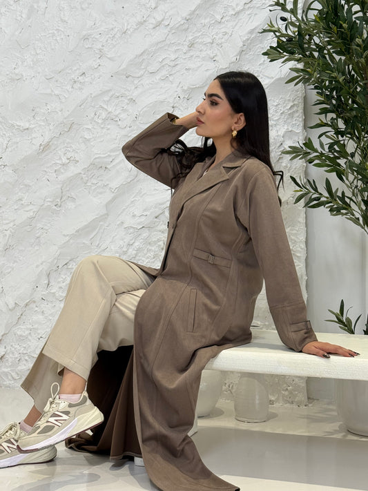 Woman in a Light Brown Shamwa Trench Abaya Set standing against a white textured wall with greenery.