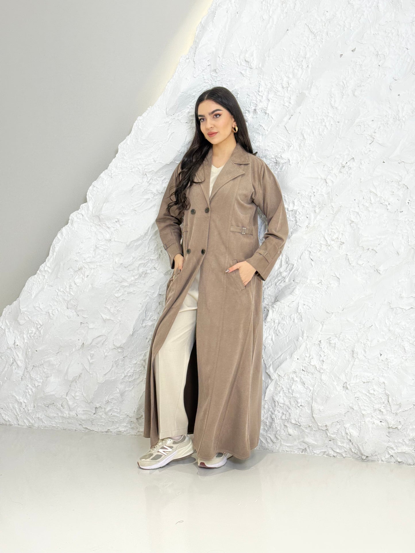 Woman in a Light Brown Shamwa Trench Abaya Sett standing against a textured white wall.