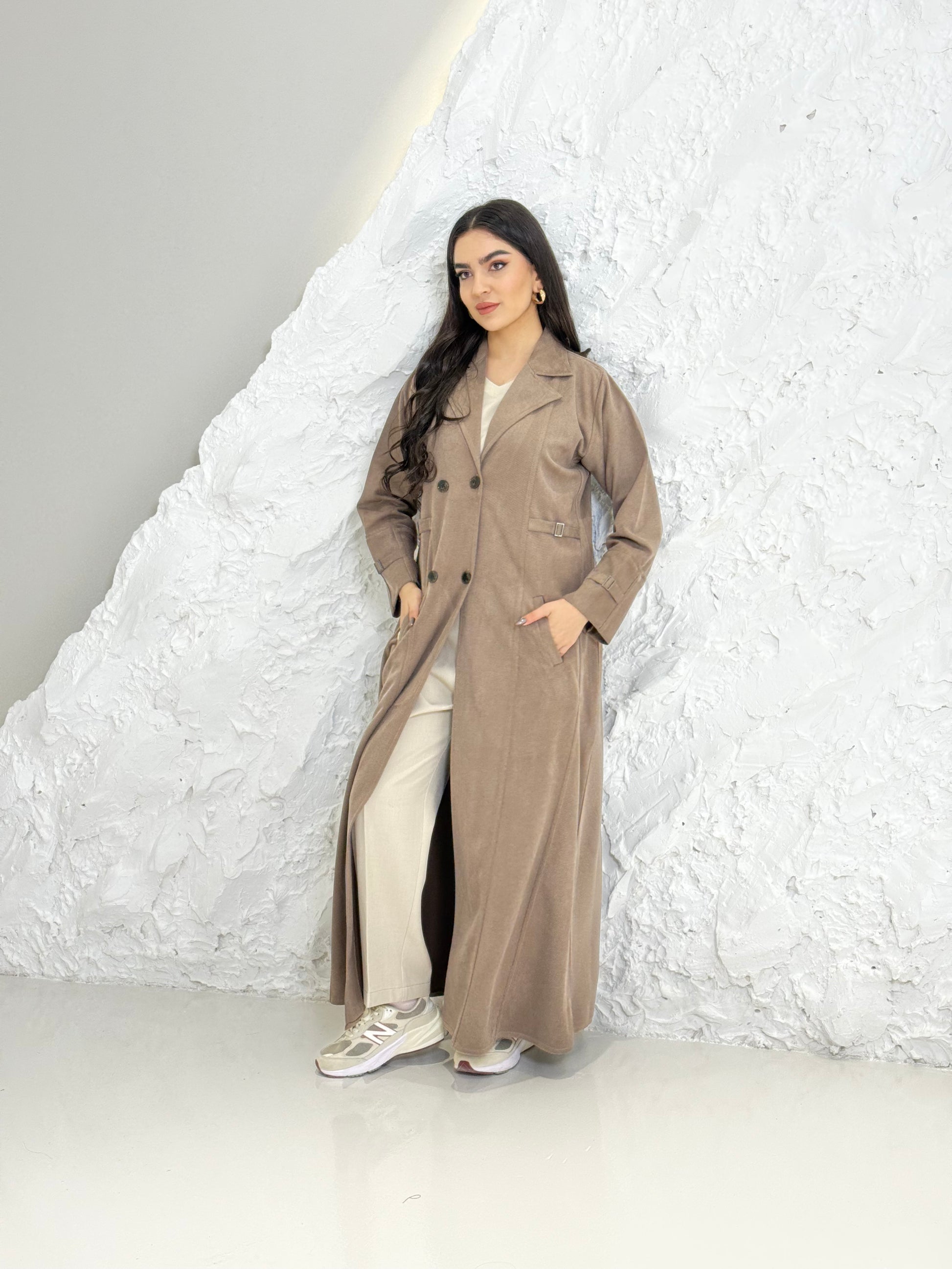 Woman in a Light Brown Shamwa Trench Abaya Sett standing against a textured white wall.