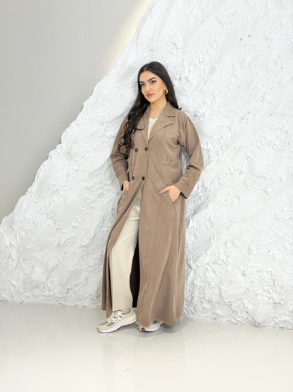 Woman in a Light Brown Shamwa Trench Abaya Sett standing against a textured white wall.