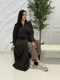 Person wearing a long Dark olive Linen Abaya trench coat Set sitting in front of a white door with gold handles.