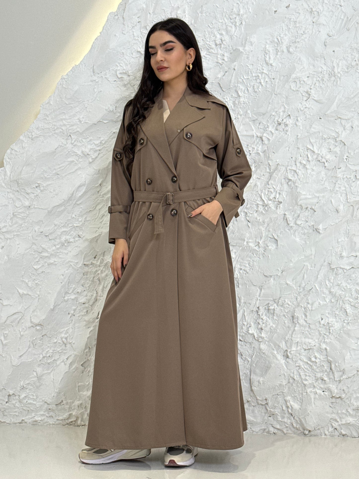 Person wearing a long light brownLinen Abaya trench coat Set standing in front of a white door with gold handles.