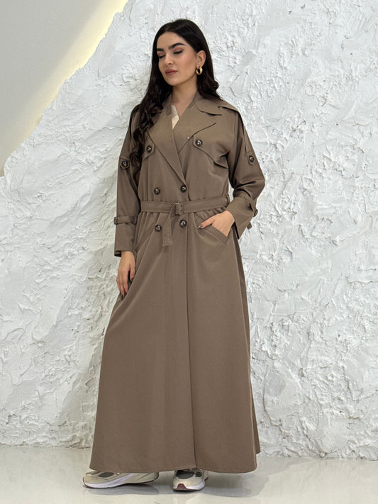 Person wearing a long light brownLinen Abaya trench coat Set standing in front of a white door with gold handles.