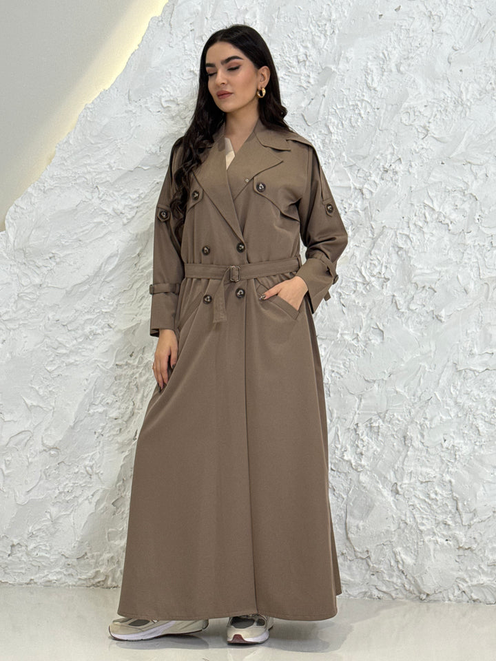 Person wearing a long light brownLinen Abaya trench coat Set standing in front of a white door with gold handles.