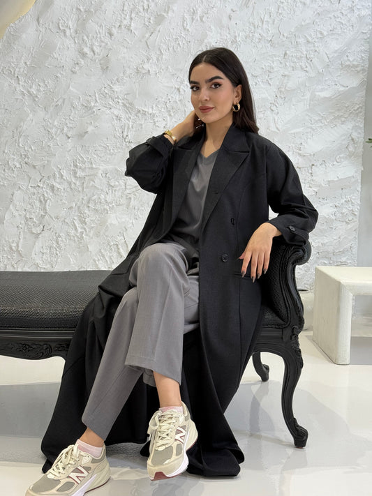 Woman in a Black Jookh Abaya Set sitting on a white bench with a textured white wall background